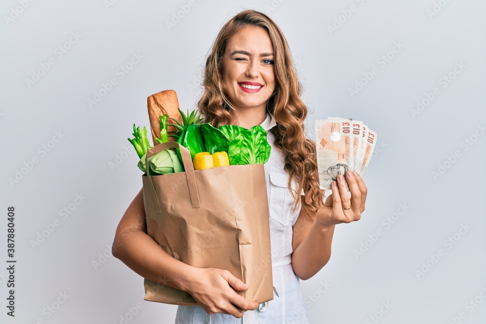 Young blonde girl holding paper bag with groceries and pounds winking looking at the camera with sexy expression, cheerful and happy face. Young blonde girl holding paper bag with groceries and pounds winking looking at the camera with sexy expression, cheerful and happy face.