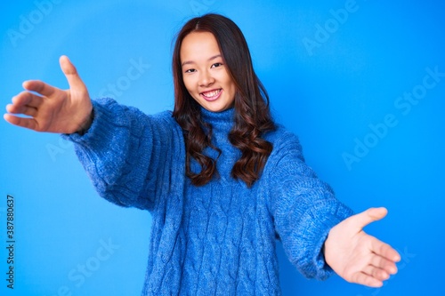 Young beautiful chinese girl wearing casual winter sweater looking at the camera smiling with open arms for hug. cheerful expression embracing happiness.