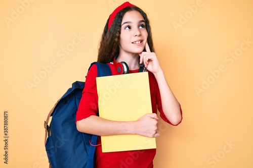 Cute hispanic child girl wearing student backpack and headphones holding book serious face thinking about question with hand on chin, thoughtful about confusing idea