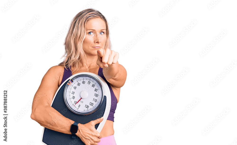 Middle age fit blonde woman wearing sports clothes holding weighing machine pointing with finger to the camera and to you, confident gesture looking serious Middle age fit blonde woman wearing sports clothes holding weighing machine pointing with finger to the camera and to you, confident gesture looking serious