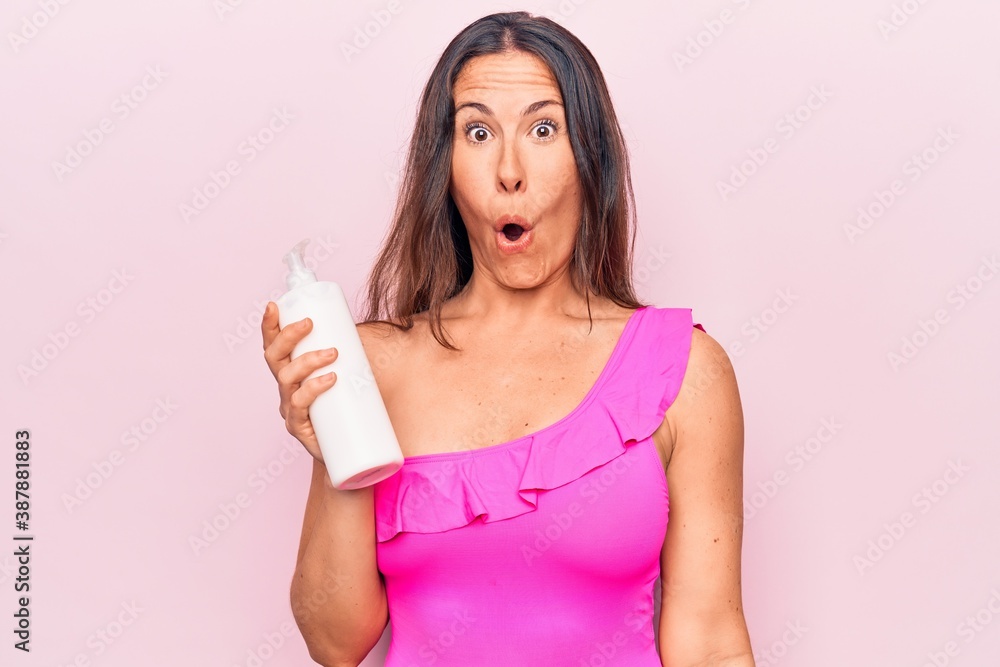 Young beautiful brunette woman wearing pink swimwear holding bottle of sunscreen lotion scared and amazed with open mouth for surprise, disbelief face Young beautiful brunette woman wearing pink swimwear holding bottle of sunscreen lotion scared and amazed with open mouth for surprise, disbelief face