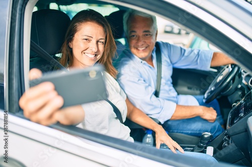 Middle age couple in love sitting inside the car going for a trip taking a selfie picture with smartphone smiling happy and cheerful together