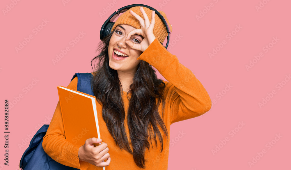 Beautiful brunette young woman wearing student backpack and holding books smiling happy doing ok sign with hand on eye looking through fingers Beautiful brunette young woman wearing student backpack and holding books smiling happy doing ok sign with hand on eye looking through fingers