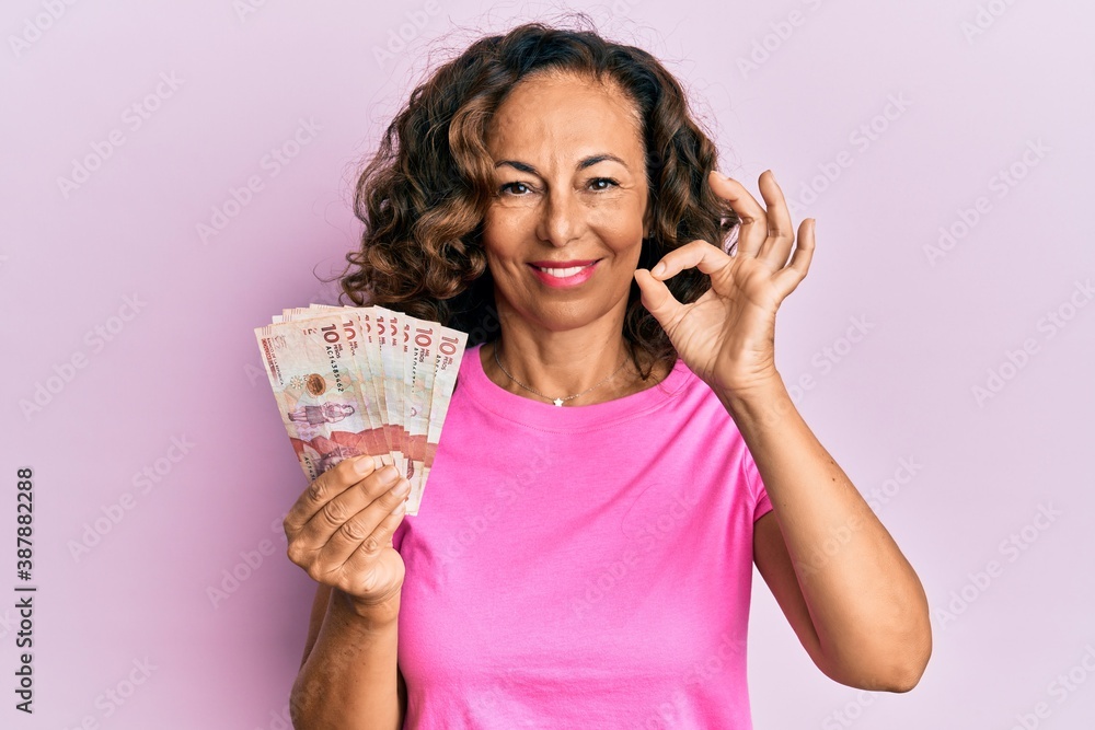 Middle age hispanic woman holding 10 colombian pesos banknotes doing ok sign with fingers, smiling friendly gesturing excellent symbol Middle age hispanic woman holding 10 colombian pesos banknotes doing ok sign with fingers, smiling friendly gesturing excellent symbol