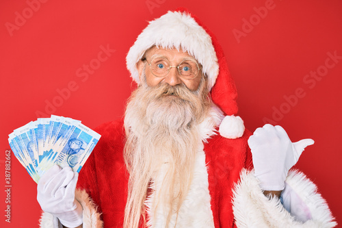 Old senior man with grey hair and long beard wearing santa claus costume holding african rands pointing thumb up to the side smiling happy with open mouth