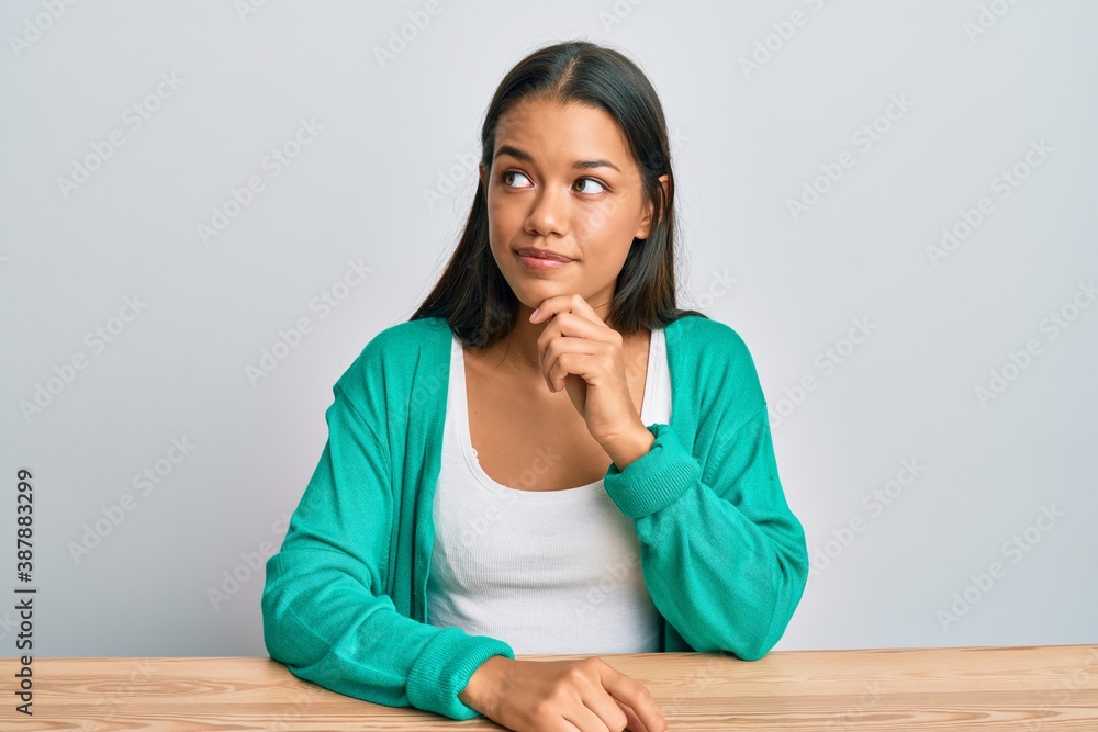 Beautiful hispanic woman wearing casual clothes sitting on the table thinking concentrated about doubt with finger on chin and looking up wondering Beautiful hispanic woman wearing casual clothes sitting on the table thinking concentrated about doubt with finger on chin and looking up wondering