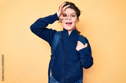 Cute hispanic child holding student backpack smiling happy doing ok sign with hand on eye looking through fingers