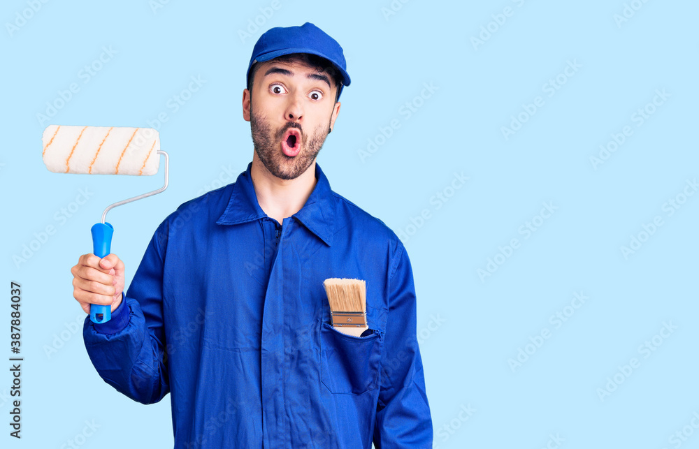 Young hispanic man wearing painter uniform holding roller scared and amazed with open mouth for surprise, disbelief face Young hispanic man wearing painter uniform holding roller scared and amazed with open mouth for surprise, disbelief face