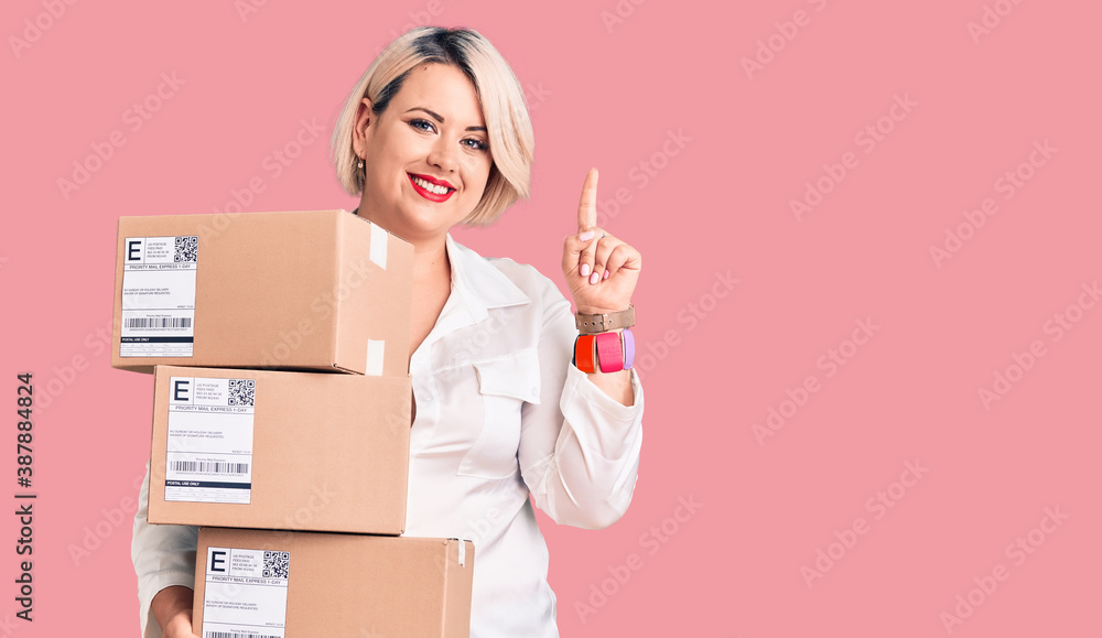 Young blonde plus size woman holding delivery cardboard package surprised with an idea or question pointing finger with happy face, number one Young blonde plus size woman holding delivery cardboard package surprised with an idea or question pointing finger with happy face, number one
