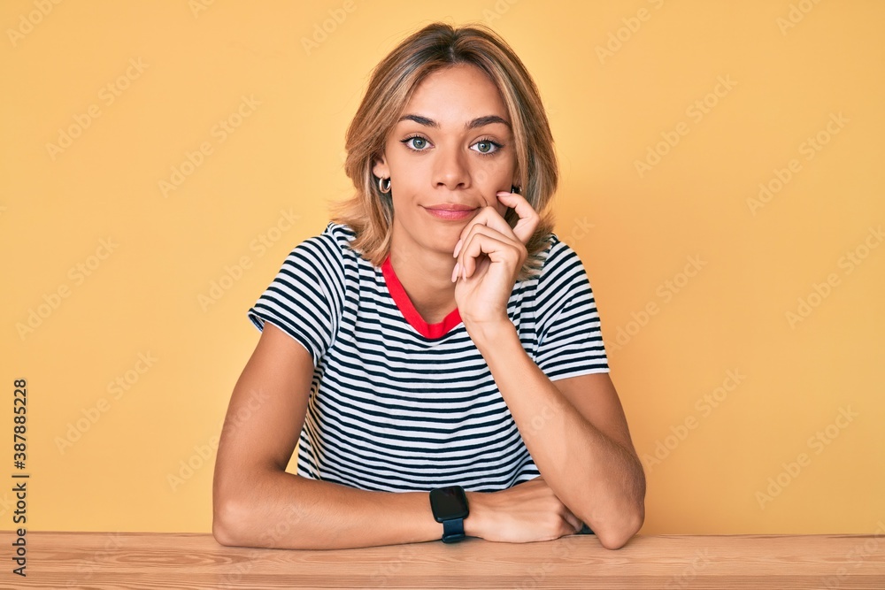 Beautiful caucasian woman wearing casual clothes sitting on the table serious face thinking about question with hand on chin, thoughtful about confusing idea