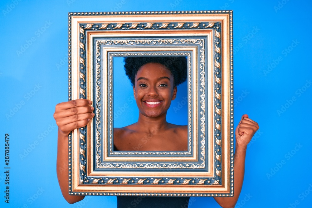 Obraz premium Young african american woman holding empty frame screaming proud, celebrating victory and success very excited with raised arm