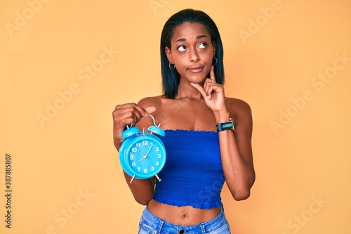 Young african american girl holding alarm clock serious face thinking about question with hand on chin, thoughtful about confusing idea