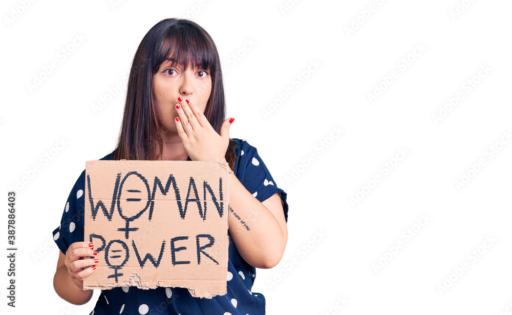 Young plus size woman holding woman power banner covering mouth with hand, shocked and afraid for mistake. surprised expression Young plus size woman holding woman power banner covering mouth with hand, shocked and afraid for mistake. surprised expression