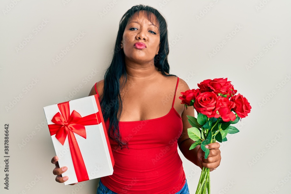 Middle age african american woman holding anniversary present and bouquet of flowers looking at the camera blowing a kiss being lovely and sexy. love expression. Middle age african american woman holding anniversary present and bouquet of flowers looking at the camera blowing a kiss being lovely and sexy. love expression.