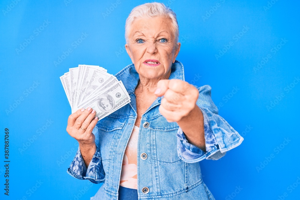 Senior beautiful woman with blue eyes and grey hair holding dollars annoyed and frustrated shouting with anger, yelling crazy with anger and hand raised Senior beautiful woman with blue eyes and grey hair holding dollars annoyed and frustrated shouting with anger, yelling crazy with anger and hand raised