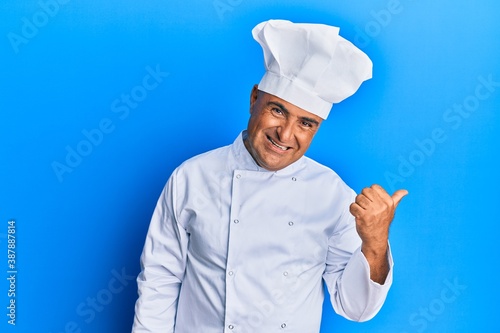 Mature middle east man wearing professional cook uniform and hat smiling with happy face looking and pointing to the side with thumb up.
