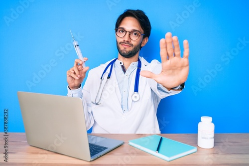 Handsome hispanic doctor man at the clinic holding syringe with open hand doing stop sign with serious and confident expression  defense gesture