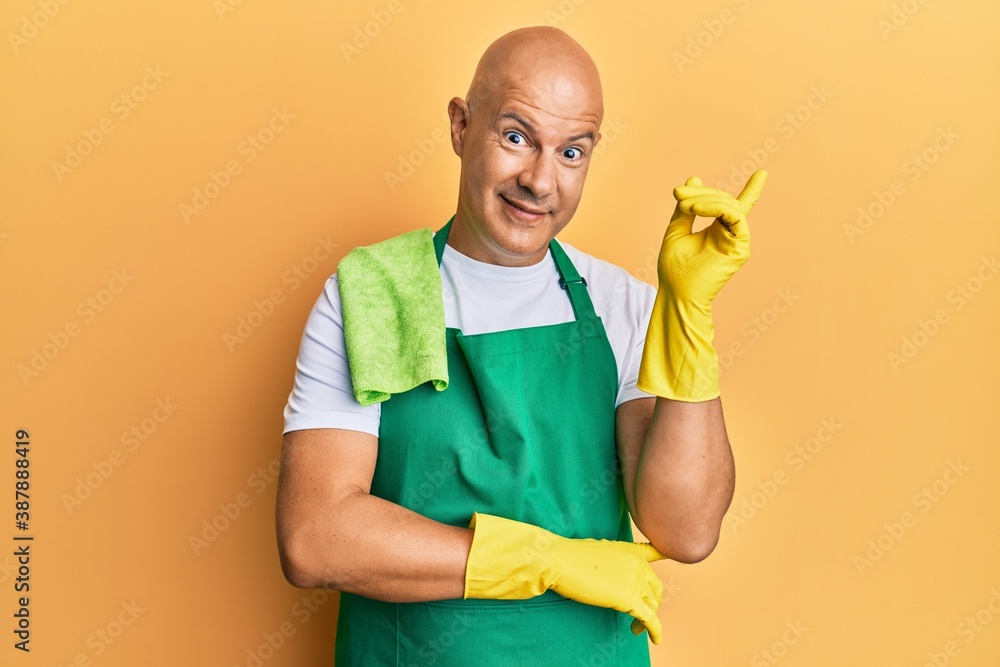 Middle age bald man wearing apron holding cleaning gloves smiling happy pointing with hand and finger to the side Middle age bald man wearing apron holding cleaning gloves smiling happy pointing with hand and finger to the side