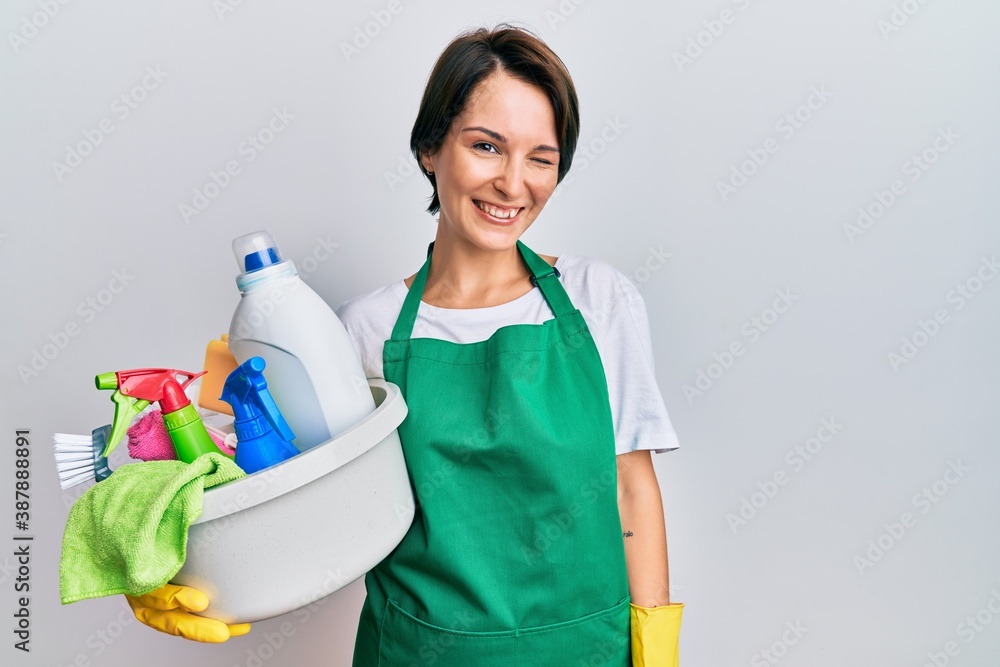 Young brunette woman with short hair wearing apron holding cleaning products winking looking at the camera with sexy expression, cheerful and happy face. Young brunette woman with short hair wearing apron holding cleaning products winking looking at the camera with sexy expression, cheerful and happy face.