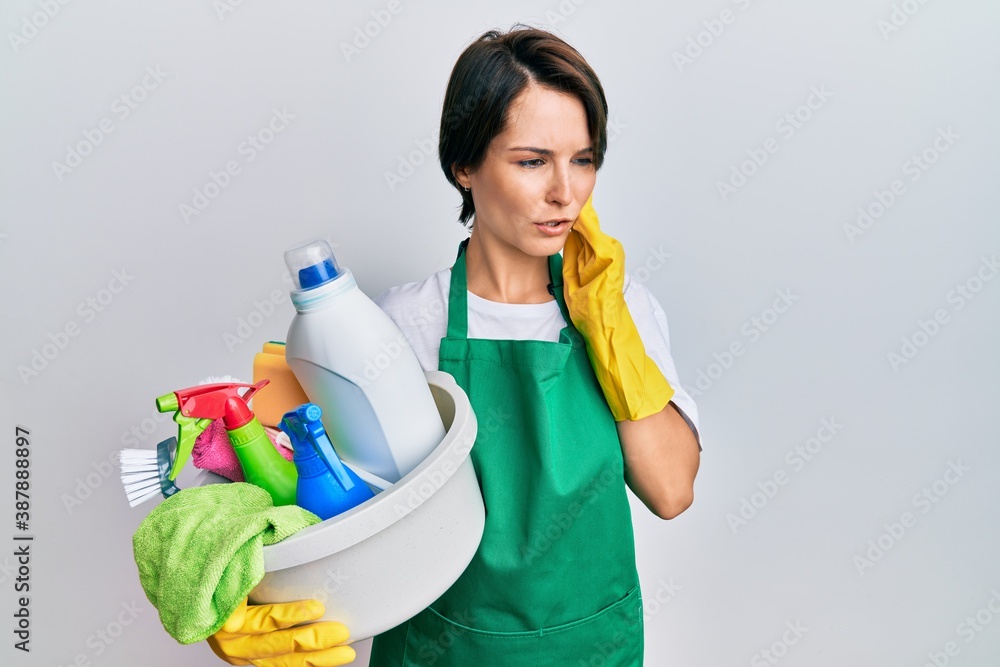 Young brunette woman with short hair wearing apron holding cleaning products touching mouth with hand with painful expression because of toothache or dental illness on teeth. dentist concept. Young brunette woman with short hair wearing apron holding cleaning products touching mouth with hand with painful expression because of toothache or dental illness on teeth. dentist concept.
