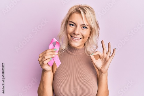 Young blonde girl holding pink cancer ribbon doing ok sign with fingers, smiling friendly gesturing excellent symbol