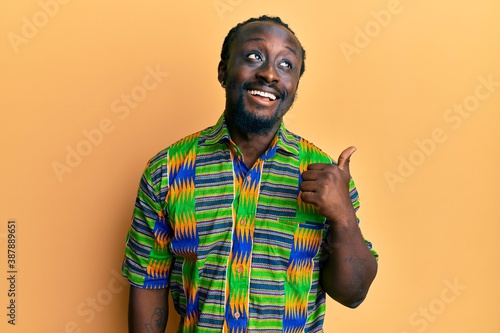 Handsome young black man wearing colorful ethnic clothes pointing thumb up to the side smiling happy with open mouth