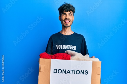 Young hispanic man holding donation box with clothes sticking tongue out happy with funny expression.