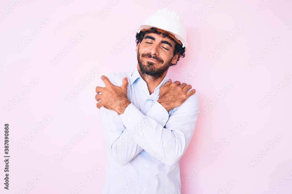 Handsome young man with curly hair and bear wearing architect hardhat hugging oneself happy and positive, smiling confident. self love and self care Handsome young man with curly hair and bear wearing architect hardhat hugging oneself happy and positive, smiling confident. self love and self care