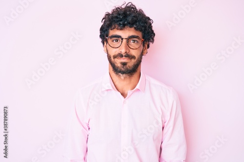 Handsome young man with curly hair and bear wearing business shirt and glasses with serious expression on face. simple and natural looking at the camera.