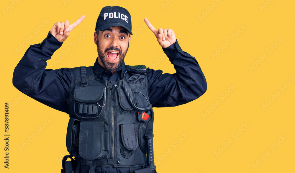 Young hispanic man wearing police uniform smiling amazed and surprised and pointing up with fingers and raised arms.