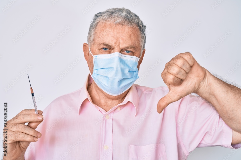 Senior grey-haired man wearing medical mask holding syringe with angry face, negative sign showing dislike with thumbs down, rejection concept Senior grey-haired man wearing medical mask holding syringe with angry face, negative sign showing dislike with thumbs down, rejection concept