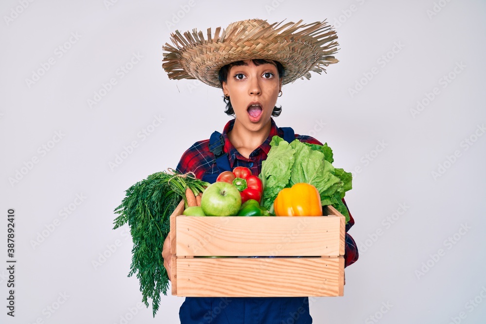 Beautiful brunettte woman wearing farmer clothes holding vegetables afraid and shocked with surprise and amazed expression, fear and excited face.