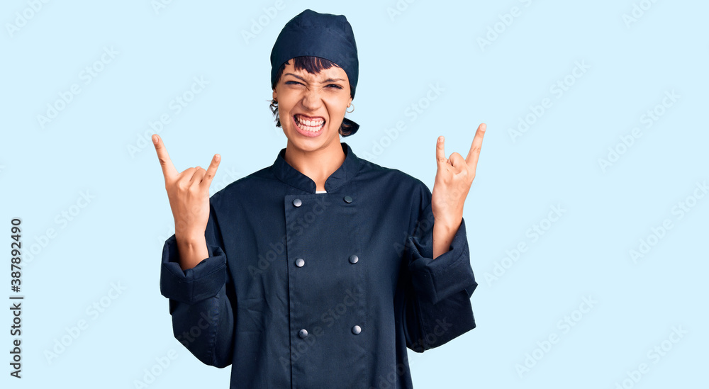 Young brunette woman with short hair wearing professional cook uniform shouting with crazy expression doing rock symbol with hands up. music star. heavy concept.