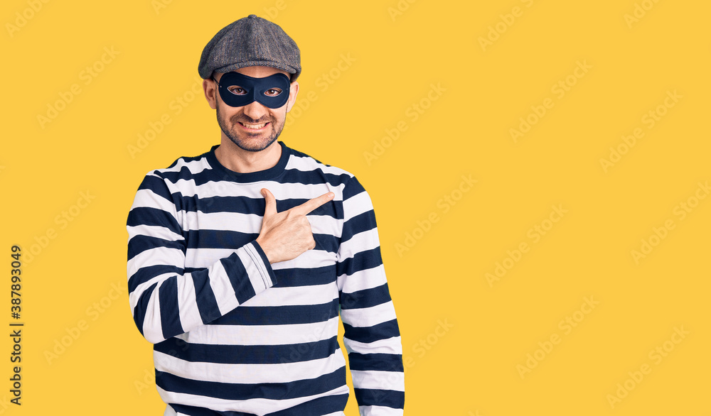 Fototapeta premium Young handsome man wearing burglar mask cheerful with a smile on face pointing with hand and finger up to the side with happy and natural expression