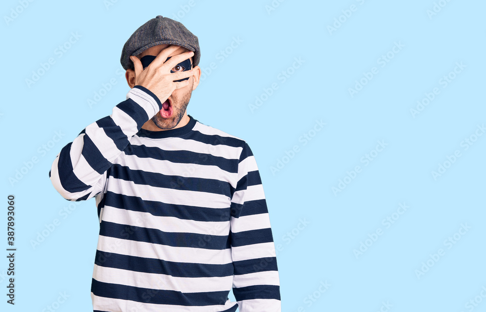 Young handsome man wearing burglar mask peeking in shock covering face and eyes with hand, looking through fingers with embarrassed expression. Young handsome man wearing burglar mask peeking in shock covering face and eyes with hand, looking through fingers with embarrassed expression.