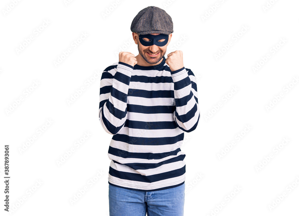 Young handsome man wearing burglar mask excited for success with arms raised and eyes closed celebrating victory smiling. winner concept. Young handsome man wearing burglar mask excited for success with arms raised and eyes closed celebrating victory smiling. winner concept.