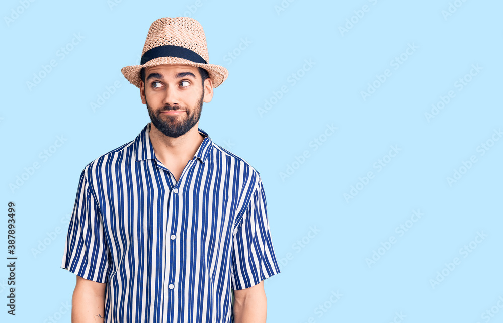 Young handsome man with beard wearing summer hat and striped shirt smiling looking to the side and staring away thinking. Young handsome man with beard wearing summer hat and striped shirt smiling looking to the side and staring away thinking.