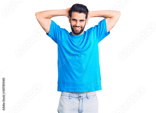 Young handsome man with beard wearing casual t-shirt relaxing and stretching, arms and hands behind head and neck smiling happy
