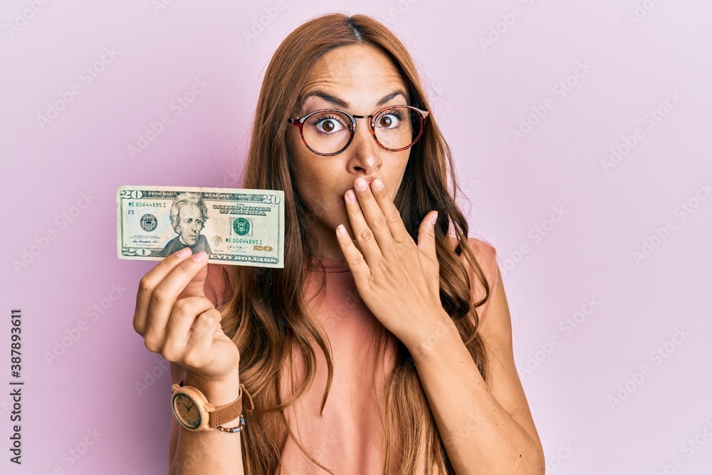 Young brunette woman holding 20 dollars banknote covering mouth with hand, shocked and afraid for mistake. surprised expression Young brunette woman holding 20 dollars banknote covering mouth with hand, shocked and afraid for mistake. surprised expression