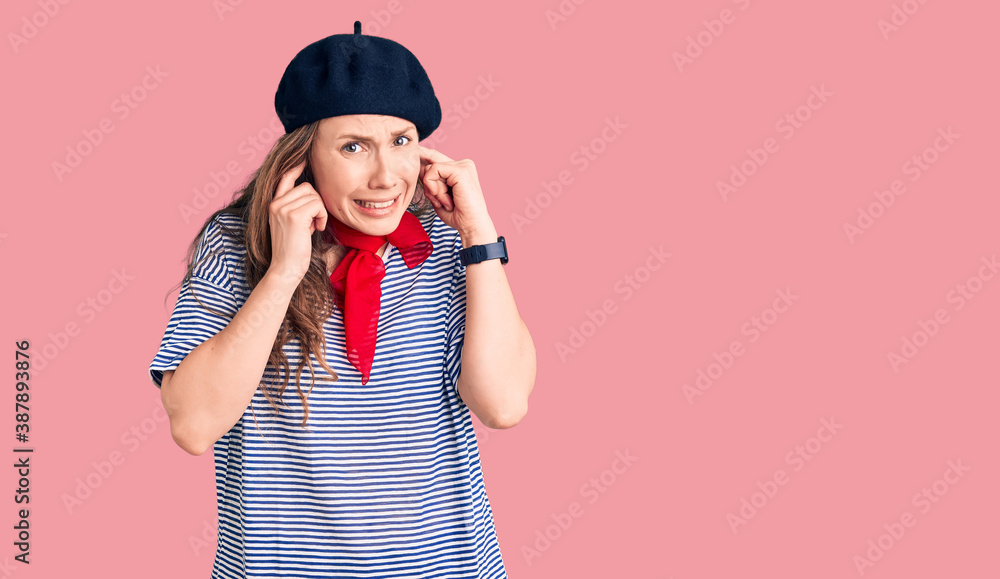 Young beautiful blonde woman wearing french beret and striped t-shirt covering ears with fingers with annoyed expression for the noise of loud music. deaf concept.