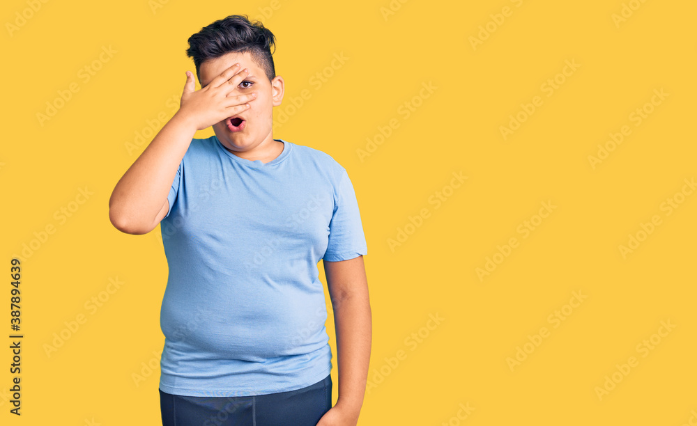 Little boy kid wearing sports workout clothes peeking in shock covering face and eyes with hand, looking through fingers with embarrassed expression. Little boy kid wearing sports workout clothes peeking in shock covering face and eyes with hand, looking through fingers with embarrassed expression.