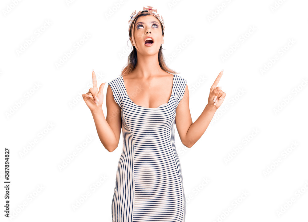 Young beautiful woman wearing king crown amazed and surprised looking up and pointing with fingers and raised arms. Young beautiful woman wearing king crown amazed and surprised looking up and pointing with fingers and raised arms.
