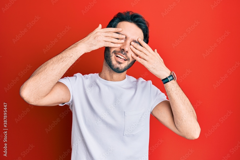 Young hispanic man wearing casual white tshirt covering eyes with hands smiling cheerful and funny. blind concept.