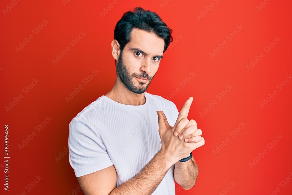 Young hispanic man wearing casual white tshirt holding symbolic gun with hand gesture, playing killing shooting weapons, angry face Young hispanic man wearing casual white tshirt holding symbolic gun with hand gesture, playing killing shooting weapons, angry face