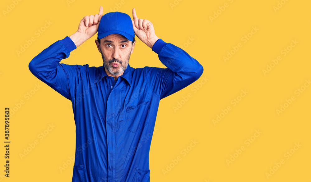 Middle age handsome man wearing mechanic uniform doing funny gesture with finger over head as bull horns