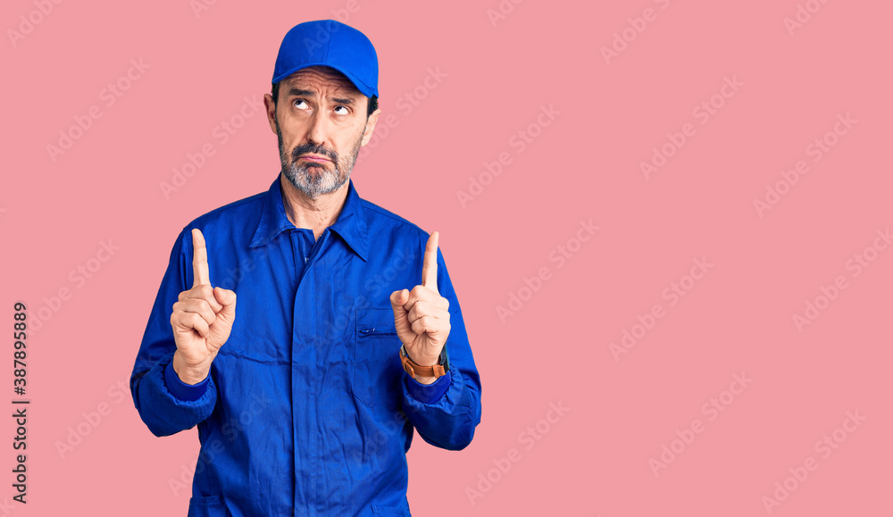 Fototapeta premium Middle age handsome man wearing mechanic uniform pointing up looking sad and upset, indicating direction with fingers, unhappy and depressed.