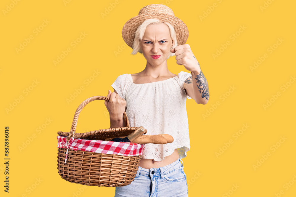 Young blonde woman with tattoo wearing summer hat and holding picnic wicker basket with bread annoyed and frustrated shouting with anger, yelling crazy with anger and hand raised Young blonde woman with tattoo wearing summer hat and holding picnic wicker basket with bread annoyed and frustrated shouting with anger, yelling crazy with anger and hand raised
