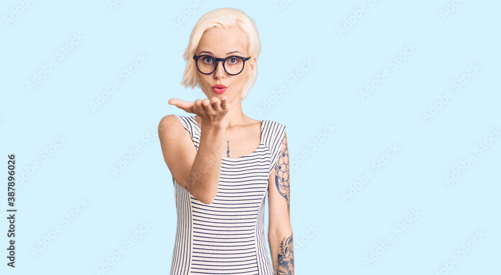 Young blonde woman with tattoo wearing casual clothes and glasses looking at the camera blowing a kiss with hand on air being lovely and sexy. love expression.