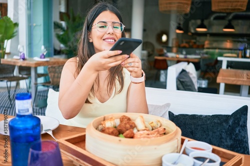 Young beautiful hispanic woman smiling happy. Sitting on the table using smartphone to make food picture at restaurant