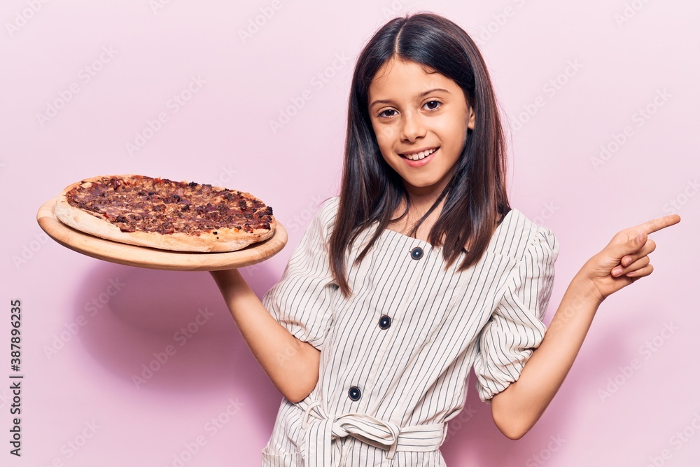 Beautiful child girl holding italian pizza smiling happy pointing with hand and finger to the side Beautiful child girl holding italian pizza smiling happy pointing with hand and finger to the side